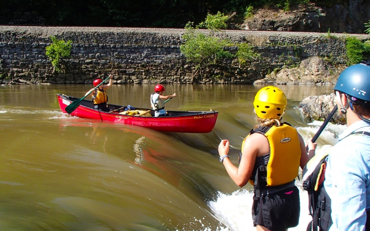 Two people wearing safety gear navigate a canoe over a short waterfall. Two more people stand on the shore, watching them.